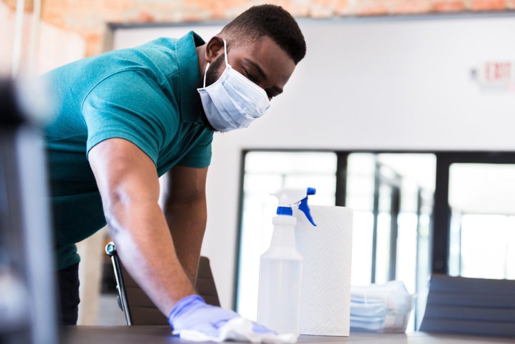 man cleans a conference table in his office while wearing a protective face mask.